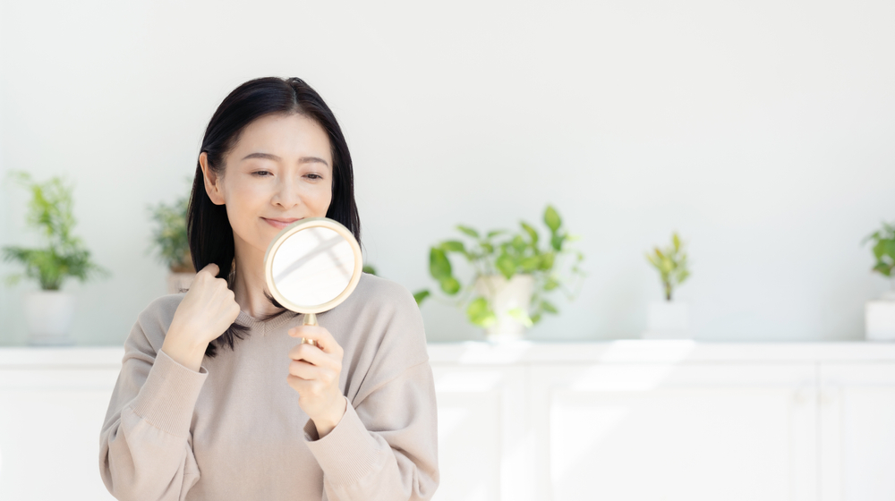 女性微笑對著鏡子，仔細檢視自己容顏，展現自信和護膚意識。